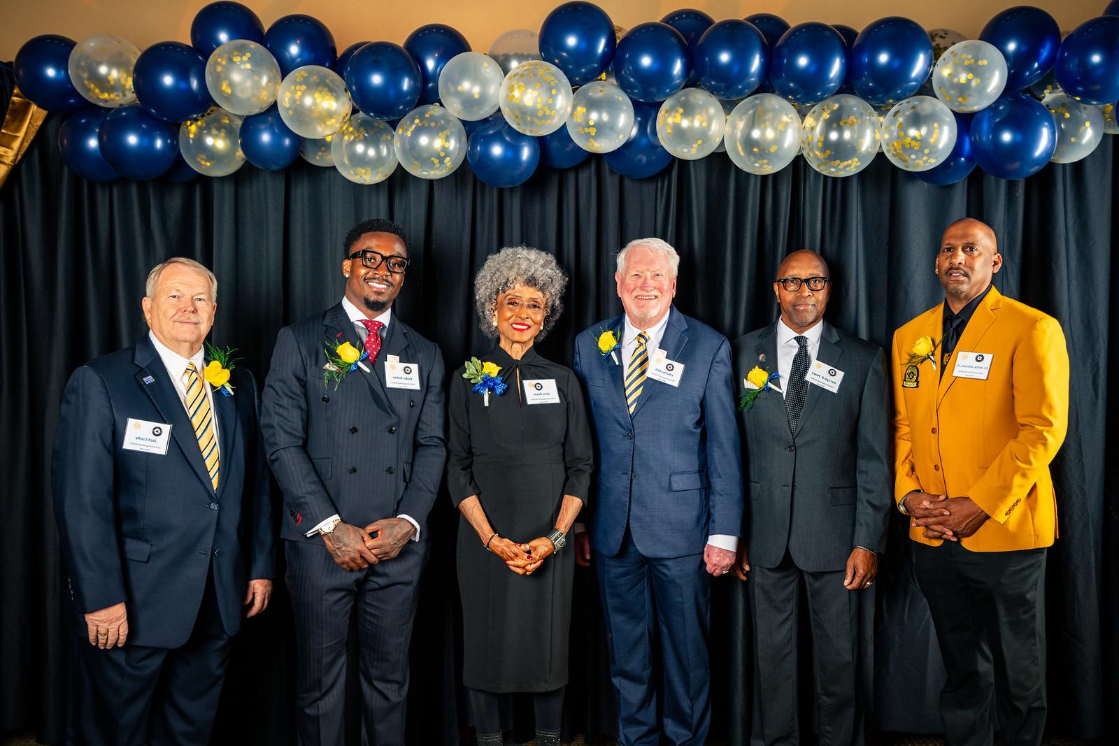 The six award recipients line up for a photo in front of a black curtain with gold and blue balloons above their heads.